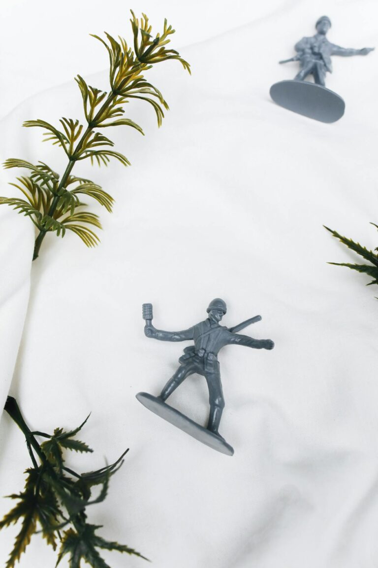 Two plastic toy soldiers on white fabric background with artificial foliage.